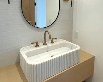 May include: A white marble bathroom sink with a fluted design and a brass faucet. The sink is mounted on a light brown countertop.