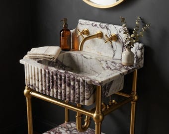 May include: A white and purple marble sink with a fluted front, supported by a brass metal frame. The sink features a brass faucet with two cross-handle knobs, a matching marble lower shelf, a brown glass soap dispenser, and a small vase with greenery.