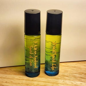 May include: Two glass roll-on bottles with black caps. The bottles have a gradient of blue, green, and yellow liquid inside. Gold text on the left bottle reads "I Am an Awakened Spark of Love." The right bottle reads "Awakened Spark. Life."