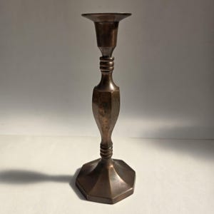 May include: A tall, metallic candlestick holder with a dark bronze finish. It features a wide, flat circular top, a slender neck with two sets of horizontal ringed accents, a central hexagonal body, and a wide, stable hexagonal base.