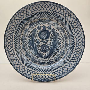 May include: A round ceramic plate featuring a hand-painted dark blue pattern on a light gray background. The center displays a pomegranate and floral motif, surrounded by a border of intricate lattice and diagonal line designs.