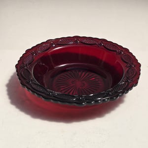 May include: A deep red glass bowl featuring a scalloped edge and a decorative rim with an embossed oval and crosshatch pattern. The center of the bowl displays a radiating sunburst design that extends outward from the middle to the smooth basin.