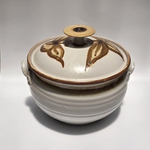 May include: A round ceramic casserole dish with a lid, featuring a speckled off-white glaze and brown hand-painted leaf motifs. The pot has two side handles and a central knob on the lid, with horizontal ridges circling the body and brown trim.