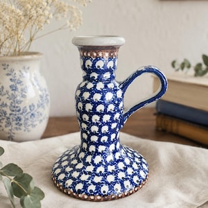 May include: A blue and white ceramic candlestick holder with a handle. The holder features a repeating pattern of white dots on a blue background. The base and rim have a brown border. A small vase with flowers is in the background.