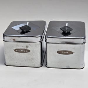 May include: Two square, chrome-finished metal canisters with rounded corners. Each canister features a black, diamond-shaped knob on the lid and an oval label on the front. One label reads coffee and the other label reads tea in white lettering.