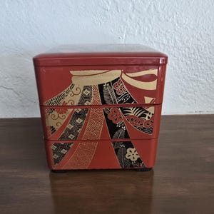 May include: A red, three-tiered Japanese bento box with gold and black decorative patterns. The box has a rectangular shape and is made of lacquered wood. The design includes floral and geometric motifs.