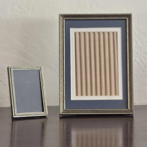 May include: Two rectangular picture frames with metallic silver-toned borders. The smaller frame features a plain dark gray insert. The larger frame includes a dark blue matting surrounding a central beige panel with a vertical ribbed texture.