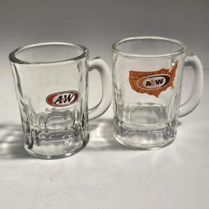 May include: Two clear glass mugs with handles. The left mug features the A&W logo with the text All American Food. The right mug displays an orange map of the United States with the A&W logo centered over the map. Both mugs have a dimpled base.
