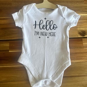 May include: White baby bodysuit with short sleeves and a snap closure. The text "Hello I'm New Here" is printed in black script with star accents. The bodysuit is hanging on a wooden hanger.