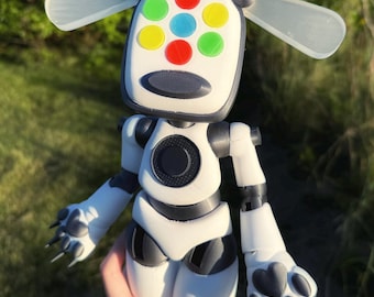 May include: A white and black robot figure with a rectangular head featuring colorful circular accents. The robot has articulated limbs, paw-like hands, and transparent ear-like extensions. The figure is held in a person's hand.