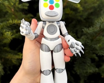 May include: A white and gray robot figurine with a square head featuring colorful circular accents. The robot has articulated limbs and claw-like hands. The figure is held in a person's hand against a blurred green background.