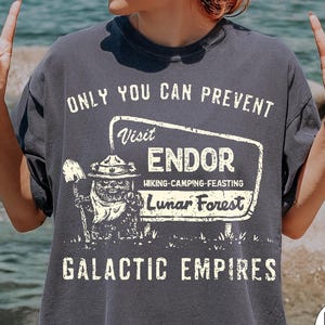 May include: Dark grey t-shirt with the text "ONLY YOU CAN PREVENT" and "Visit ENDOR Lunar Forest". The shirt features a graphic of a creature with a shovel. The text "GALACTIC EMPIRES" is also printed on the shirt.