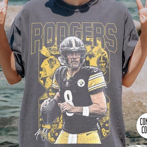 May include: A grey Comfort Colors t-shirt with a football player design. The name "RODGERS" is printed in yellow block letters. The player wears a black and yellow uniform with the number 8. The shirt also has the text "Pepper".