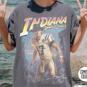 May include: Pepper-colored t-shirt featuring a graphic of a football player in a cowboy hat with the text "Indiana Daniel Jones." The shirt has a vintage, distressed look with the Comfort Colors logo.
