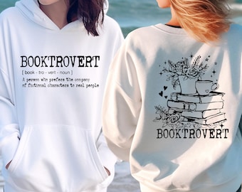 Puede incluir: Sudadera con capucha blanca con el texto "BOOKTROVERT" y una definición de la palabra. La parte posterior de la sudadera tiene una ilustración en blanco y negro de una pila de libros con flores y una taza de café.