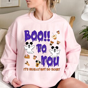 Puede incluir: Sudadera rosa claro con un diseño temático de Halloween. El diseño incluye las palabras "Boo!! To You" en letras moradas goteando, junto con fantasmas de dibujos animados y gráficos de comida. La frase "It's Really Not So Scary" está impresa debajo.