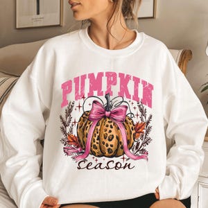 May include: White sweatshirt with a graphic of a leopard-print pumpkin adorned with a pink bow and the words "PUMPKIN SEASON" in pink. The design includes fall leaves and decorative elements, perfect for autumn.