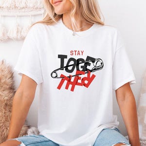 May include: White t-shirt with the words "STAY TOGETHER" in black and red, featuring a safety pin graphic. The shirt is a casual style, suitable for everyday wear.