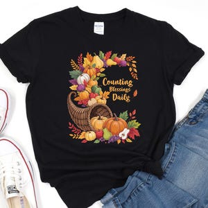 May include: Black t-shirt featuring a colorful harvest-themed graphic. The design includes a cornucopia overflowing with pumpkins, gourds, and fruit, surrounded by autumn leaves. Text reads "Counting Blessings Daily".
