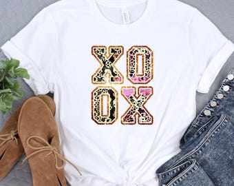 May include: White t-shirt with a graphic of the letters XOXO in a gold and pink leopard print.