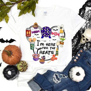 Puede incluir: Camiseta blanca con un gráfico colorido que dice "I'm here for the treats". El diseño incluye golosinas de dibujos animados, una esfera morada y azul y golosinas con temática de Mickey Mouse. La camiseta se combina con vaqueros azules, calabazas y decoraciones de Halloween.