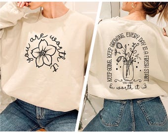 May include: A beige sweatshirt with a black floral design on the front and the text "you are worth it" with a floral design on the back.