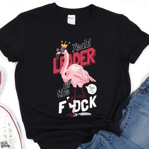 Könnte beinhalten: Schwarzes T-Shirt mit einem rosa Flamingo, der eine Sonnenbrille und eine Krone trägt. Der Flamingo ist mit "Leader of the F*ck" beschriftet, darüber steht der Text "Yeah!". Eine Sprechblase sagt "Yep! It's me."