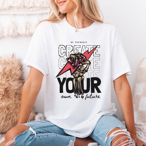May include: White t-shirt with a graphic design that reads "CREATE YOUR own future." The design includes a skeleton hand holding a red lightning bolt. The shirt is a short-sleeved crew neck.