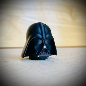 May include: A small, matte black 3D-printed figurine of a helmeted character head. The helmet features a smooth, rounded dome, a triangular face mask with a vertical grille, and two small silver-colored accents on the lower chin area.