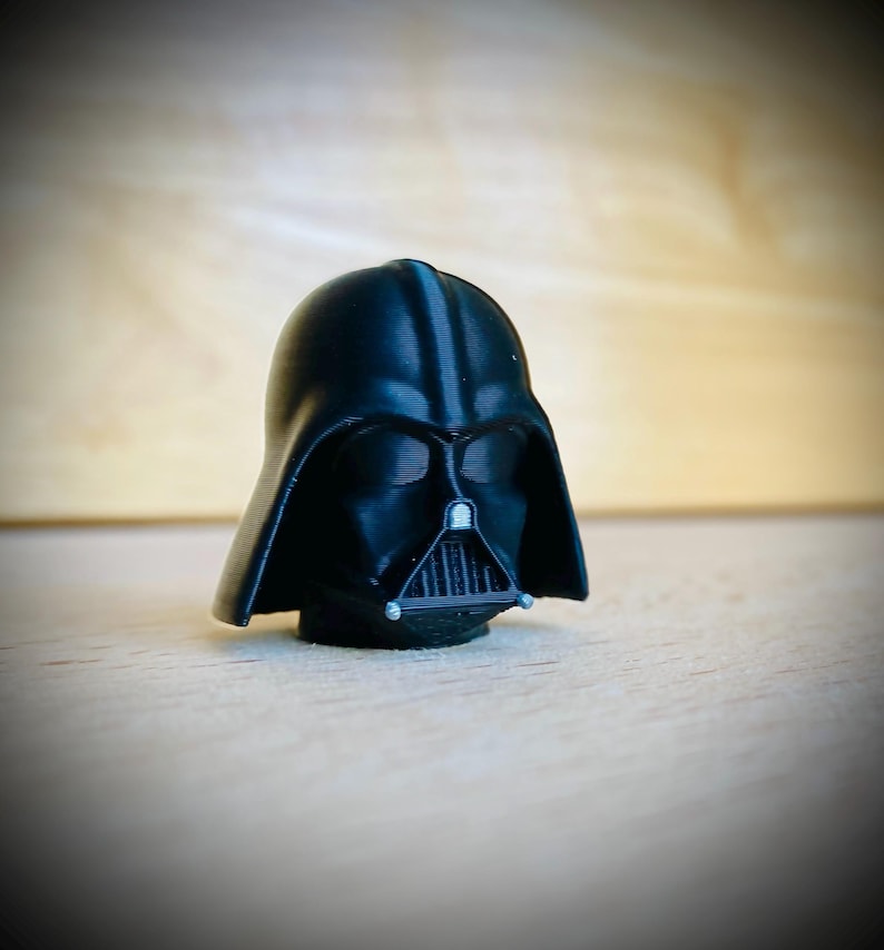 May include: A small, matte black 3D-printed figurine of a helmeted character head. The helmet features a smooth, rounded dome, a triangular face mask with a vertical grille, and two small silver-colored accents on the lower chin area.