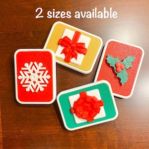 May include: Four Christmas-themed decorative items. One red rectangle with a white snowflake, one gold rectangle with a white gift and red bow, one red rectangle with green holly and red berries, and one green rectangle with a white gift and red bow. Text reads "2 sizes available".