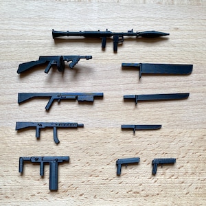 May include: A collection of black toy weapons, including a rocket launcher, submachine guns, knives, and pistols. The items are arranged on a light brown wooden surface. The collection showcases various firearm and blade designs.
