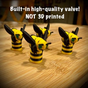 May include: Four small bee-shaped figures with yellow and black striped bodies, black wings, and green eyes. The text above reads: Built-in high-quality valve! NOT 3D printed. These figures feature a cylindrical base and detailed insect features.