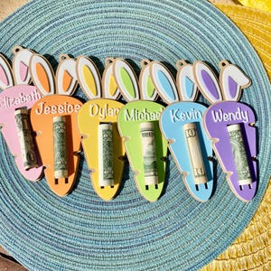 May include: Six carrot-shaped wooden ornaments in pink, orange, yellow, green, blue, and purple. Each features white bunny ears, a name engraved in white text, and a rolled-up one-dollar bill inserted into a vertical slot on the front.