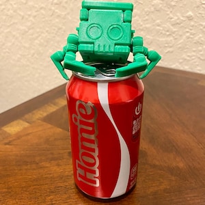 Op de afbeelding: Een rood en wit drankblikje met het woord "Homie" erop gedrukt, bekroond met een groene 3D-geprinte robotkop en armen. De robotkop heeft twee cirkelvormige oogkassen en een vierkante kop.