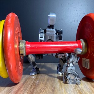 May include: Two silver robots are lifting a red barbell with black and yellow weight plates. The barbell is on a wooden surface.