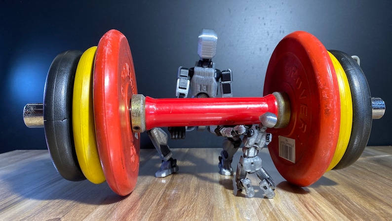 May include: Two silver robots are lifting a red barbell with black and yellow weight plates. The barbell is on a wooden surface.