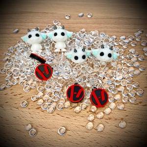 May include: White goat-like figurines and earrings with black eyes and horns. The earrings have a black and red circular design. The items are displayed on a bed of clear, round beads, with a wooden background.