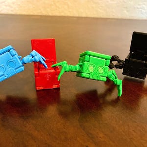 May include: Three small, colorful, articulated robot toys with spindly legs. The robots are red, green, and black. The red robot is sitting down, the green robot is standing with its legs spread out, and the black robot is standing with its legs together.