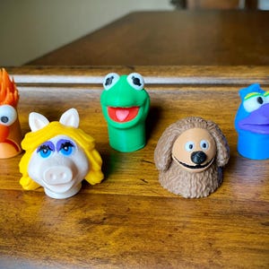Puede incluir: Un conjunto de cinco marionetas de dedo de los Muppets que incluye un personaje naranja con pelo puntiagudo, un cerdo rubio, una rana verde, un perro marrón peludo y un personaje azul con una nariz grande, todos hechos de plástico moldeado.