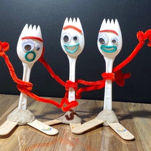 May include: Three white plastic toy forks with red pipe cleaner arms and googly eyes. The forks are arranged in a line with the middle fork in the center. The forks are wearing red pipe cleaner arms and have googly eyes. The forks are on a wooden surface.
