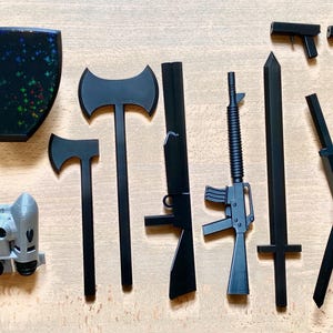 May include: A collection of black plastic weapons, including a sword, two axes, two pistols, a rifle, and two katanas. The weapons are arranged on a wooden surface.