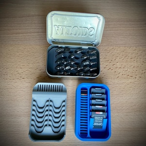 Puede incluir: Una caja metálica rectangular abierta con la palabra "VINTAGE" en relieve en la tapa, que contiene varias brocas metálicas. Debajo, un soporte gris impreso en 3D y un soporte azul impreso en 3D con tarjetas SD y una etiqueta Lexar.