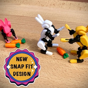 May include: Three colorful, articulated rabbit figures with carrots. One is pink, one is white and black, and one is yellow. The image also includes a sticker with the text "NEW SNAP FIT DESIGN".