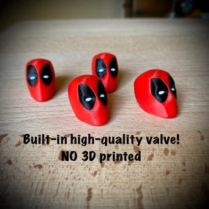 Op de afbeelding: Vier rode Deadpool-hoofdvormige objecten met zwarte oog- en monddetails en witte ogen. De tekst "Built-in high-quality valve! NO 3D printed" staat eronder. Op een houten oppervlak.