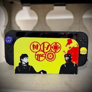 May include: Black Nintendo Switch console with a yellow and red graphic cover featuring band imagery and two black figures. The console has blue and yellow joystick caps. The cover has a band logo and a red splatter design.