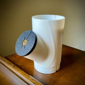 May include: A white plastic rain barrel diverter with a black and gold filter. The diverter has a cylindrical body with a side outlet and is designed to collect rainwater from a downspout. The filter is circular and has a starburst pattern.