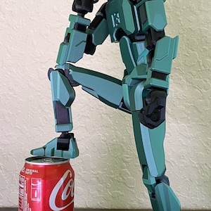 May include: A teal green robot figurine with black accents is posed in a thinking position with one leg resting on a red Coca-Cola can. The can has the words "Recycle Me" printed on it.