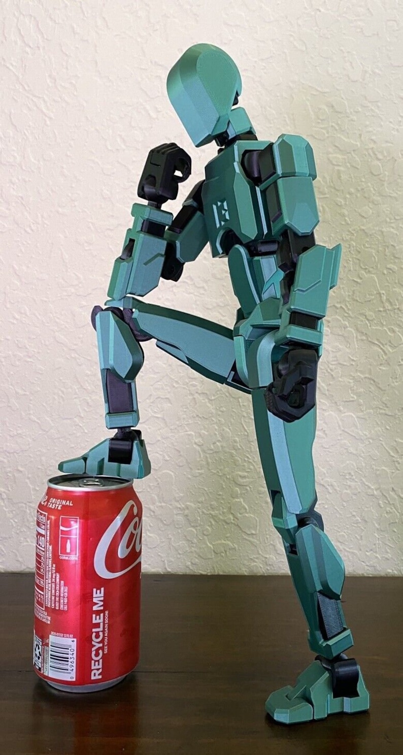 May include: A teal green robot figurine with black accents is posed in a thinking position with one leg resting on a red Coca-Cola can. The can has the words "Recycle Me" printed on it.