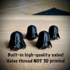 May include: Four black 3D printed helmet-shaped valve caps. The text below reads: Built-in high-quality valve! Valve thread NOT 3D printed. Each helmet features a smooth finish with a detailed face mask and a central silver-colored accent.
